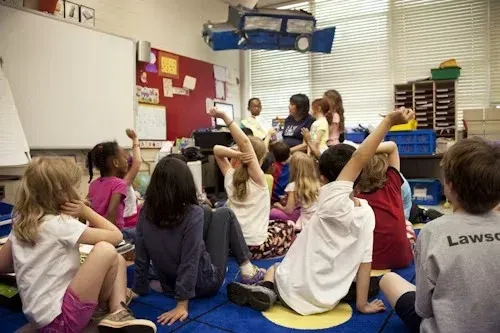 Children sitting on a carpet raising their hands to answer a question